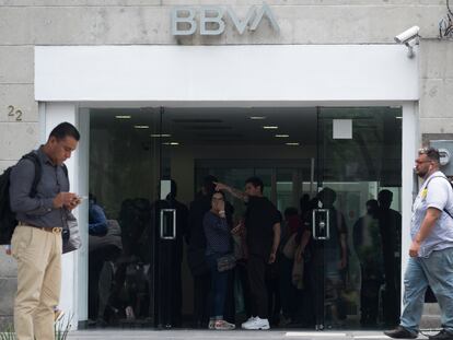 Usuarios frente a una sucursal de BBVA en Ciudad de México que revisan su celular y hacen operaciones bancarias.