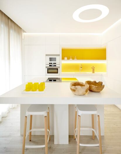 La cocina comparte espacio con el comedor y la zona de estar. Se trata de una estancia blanca con un destello de color luminoso. En el centro, una isla muy versátil hace las veces de office. Los vasos, la bandeja y los cuencos de madera que hay sobre la encimera son de la tienda de diseño Mestizo Store, en el madrileño barrio de Chueca.