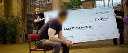 El hacker Joe Grand, a la izquierda, sostiene un cheque de 1.6 millones de dólares (valor de los bitcoin en el momento de la grabación) junto a Bruno, a la derecha, enfrente de Michael, con la cara pixelada.