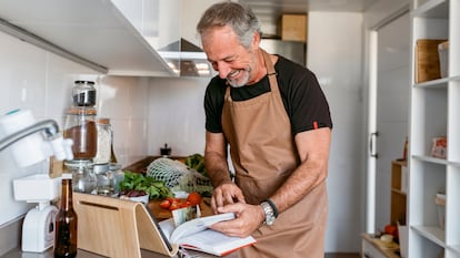 Un hombre con delantal en su cocina mirando un libro de recetas.