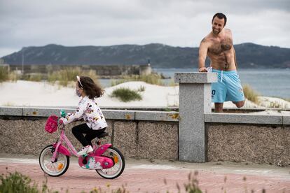 Un deportista hace estiramientos mientras una niña pasa en bicicleta por el paseo marítimo de Laxe (A Coruña).