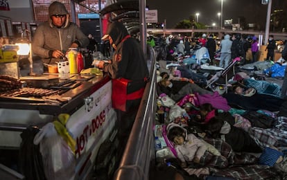 Un hombre prepara comida en un puesto en el cruce fronterizo, mientras unos migrantes se manifiestan y otros descansan en sacos de dormir sobre el suelo.