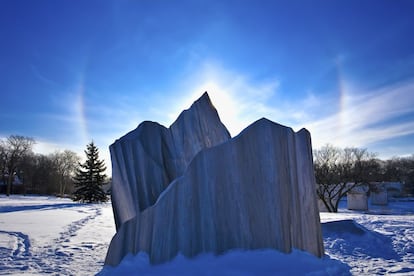 El parhelio es un efecto que se observa alrededor del sol. Son como brillantes puntos de luz (o “soles falsos”) que se localizan a ambos lados del propio astro rey. Puede durar horas. A medida que el Sol va elevándose, se van deshaciendo y toman el aspecto de cometas. Hacia el mediodía, la visión suele haber desaparecido, dejando que el verdadero sol termine solo su curso celeste. En el pasado se consideraban presagios de malos tiempos, pero, cuando se sabe que se trata únicamente de inocentes cristales de hielo que crean prismas en el aire, resultan mucho menos amenazantes. Se ven mejor si se tiene un horizonte plano y sobre todo en los amaneceres fríos o en las puestas o salidas de sol invernales. En el Ártico y en el Antártico resultan habituales. El la imagen un parhelio tras una escultura del parque natural de Assiniboine, Winnipeg, Manitoba (Canadá).