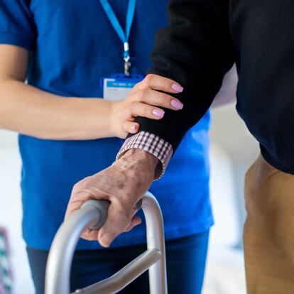Close Up Of Senior Man With Hands On Walking Frame Being Helped By Care Worker