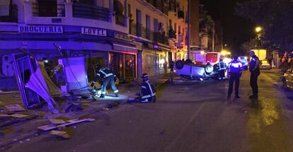 Los bomberos limpian la zona del quiosco, con el coche volcado al fondo.