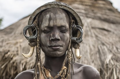 Una joven bodi de la aldea del Parque Nacional de Mago posa con los ojos cerrados y la cara pintada rápidamente con arcilla blanca y roja, con un tocado improvisado con una cacerola y el arreo de reses. El proceso de tranculturación se manifiesta no solo en la presencia de objetos autóctonos entre las decoraciones y en los poblados, sino sobre todo en la emulación y creación de decoraciones fantásticas que satisfagan a los turistas.