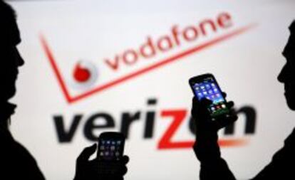 Dos usuarios miran sus m&oacute;viles ante los logotipos de Vodafone y Verizon. 