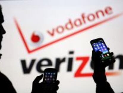 Dos usuarios miran sus m&oacute;viles ante los logotipos de Vodafone y Verizon. 