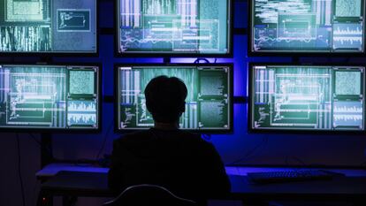Un 'hacker' con la cara cubierta en una sala con múltiples pantallas y paneles de información de seguridad de red.
