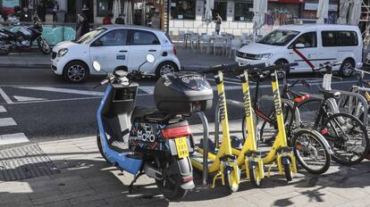 Una motocicleta y tres patinetes de alquiler por minuto aparcados en la acera. Por la calzada, un coche de una de las empresas de vehículos compartidos.