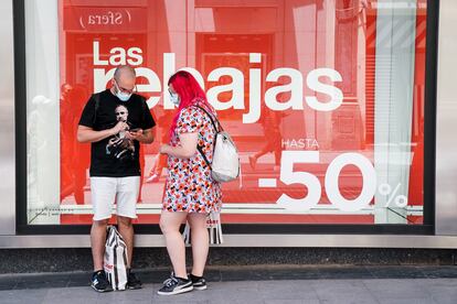 Dos personas consultan su teléfono móvil delante de un escaparate con un cartel de rebajas, el 25 de junio en Madrid.