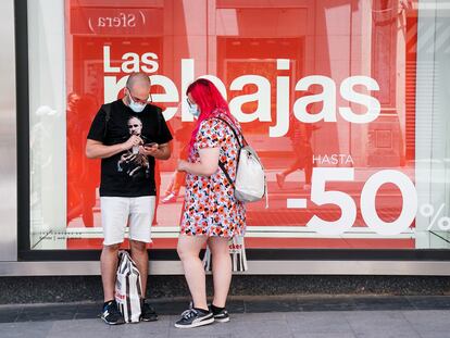 Dos personas consultan su teléfono móvil delante de un escaparate con un cartel de rebajas, el 25 de junio en Madrid.