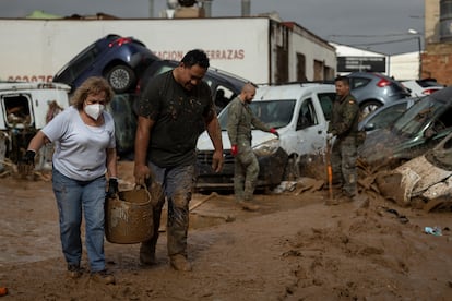 Dos personas cargan con un cubo lleno de barro, de fondo dos militares del Ejrcito de Tierra, el lunes en Paiporta (Valencia).