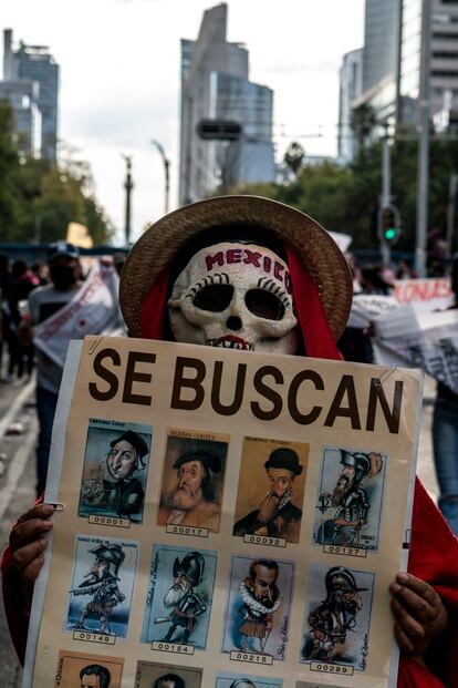 Un hombre con máscara de calavera lleva un cartel con caricaturas de conquistadores europeos como Hernán Cortés y Francisco Pizarro.