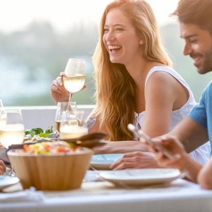 Friends chatting and drinking at a dinner party with wine glasses on the table. There are also bowls of other food and salad on the table. Time is sunset outdoors. There are 2 people using their mobile phones