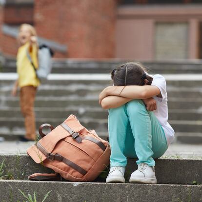 Full length portrait of crying schoolgirl sitting on stairs outdoors with group of teasing children bullying her in background, copy space