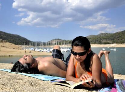 Una mujer lee un libro mientras su compañero toma el sol, en una playa del pantano de San Juan, Madrid