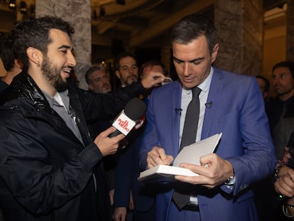 A la izquierda, Ismael Juárez, colaborador de 'El Intermedio', pasa por la espalada del presidente del Gobierno, Pedro Sánchez un décimo de la lotería de Navidad, durante la presentación del libro ‘Tierra firme’, este lunes en Madrid.