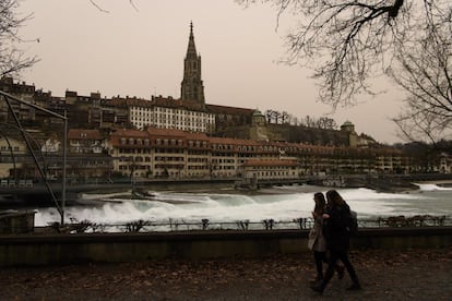 Dos mujeres caminan junto al río Aare, frente al casco antiguo y la catedral de Berna (Suiza), con el cielo coloreado de amarillo por el polvo del Sáhara.