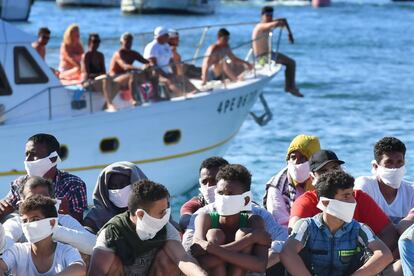 Migrantes rescatados por la Guardia Costera de Italia esperan sentados mientras un bote con turistas entra en el puerto de la isla italiana Pelagie, de Lampedusa.