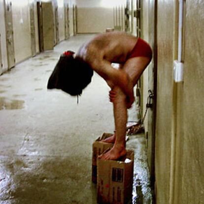 Un reo, con una capucha en la cabeza y un tobillo encadenado a una puerta, es obligado por sus captores a mantener el equilibrio sobre dos cajas de cartón en un pasillo de la prisión de Abu Ghraib.