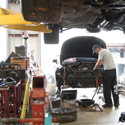 An auto repair garage.  A mechanic works on a car with it's hood up exposing the engine.  This is an active real garage, there are various tools and supplies scattered around.  In the foreground is a car up on a lift, the camera angle is underneath this car.  The mechanic is caucasian and wears a ball cap.