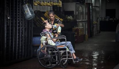 Una madre da de beber agua a su hijo postrado en una silla de ruedas en Caracas.