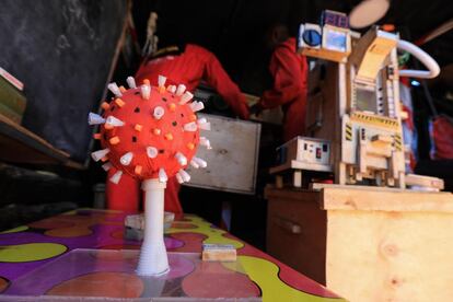 Tras una figura del virus de la covid-19, los innovadores autodidactas kenianos David Gathu (R) 29, y Moses Kiuna (L) 26, explican el funcionamiento de su 'máquina desinfectante', recientemente inventada, en su taller improvisado en Kikuyu, al norte de Nairobi, Kenia, el 26 de enero de 2021. Ni Gathu ni su socio pudieron seguir una educación superior, porque no podían asumir los costes de la universidad, y han pasado años desarrollando innovaciones tecnológicas para resolver problemas en su comunidad, desde la seguridad hasta la salud. Ahora, en medio de una pandemia, han desarrollado un dispositivo para desinfectar superficies y espacios de covid-19. Utilizando materias primas de otros aparatos desechados han innovado en los últimos años numerosas soluciones tecnológicas como un brazo robótico o una aplicación de seguridad, pero en los últimos meses han centrado sus esfuerzos en desarrollar una máquina desinfectante. El dispositivo, que aún se encuentra en fase de prototipo, oxida moléculas de oxígeno para convertirlo en otro gas. "El ozono tiene una gran capacidad como desinfectante", detalla Gathu mientras hace una demostración de su funcionamiento.