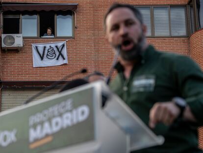 Santiago Abascal en la concentración de Vox en la Plaza de la Constitución en Vallecas, y en el fondo una vecina con una pancarta de protesta.