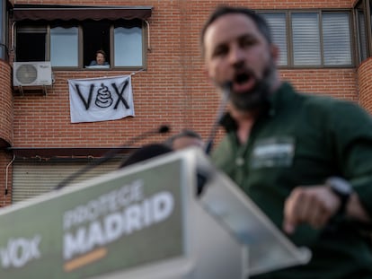 Santiago Abascal en la concentración de Vox en la Plaza de la Constitución en Vallecas, y en el fondo una vecina con una pancarta de protesta.