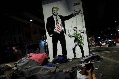 Sem-teto diante de grafite que ironiza a relação de Trump e Bolsonaro.