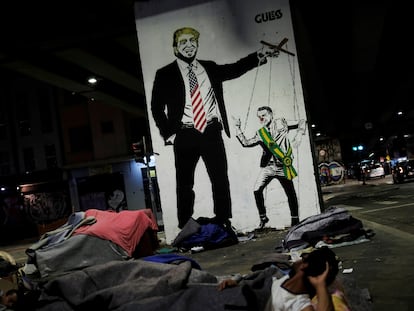 Sem-teto diante de grafite que ironiza a relação de Trump e Bolsonaro.