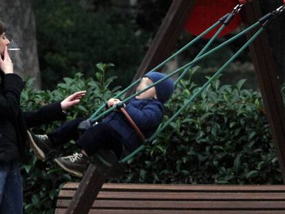 Una mujer fuma un cigarrillo ante un columpio ocupado por un niño en el parque del Retiro de Madrid.