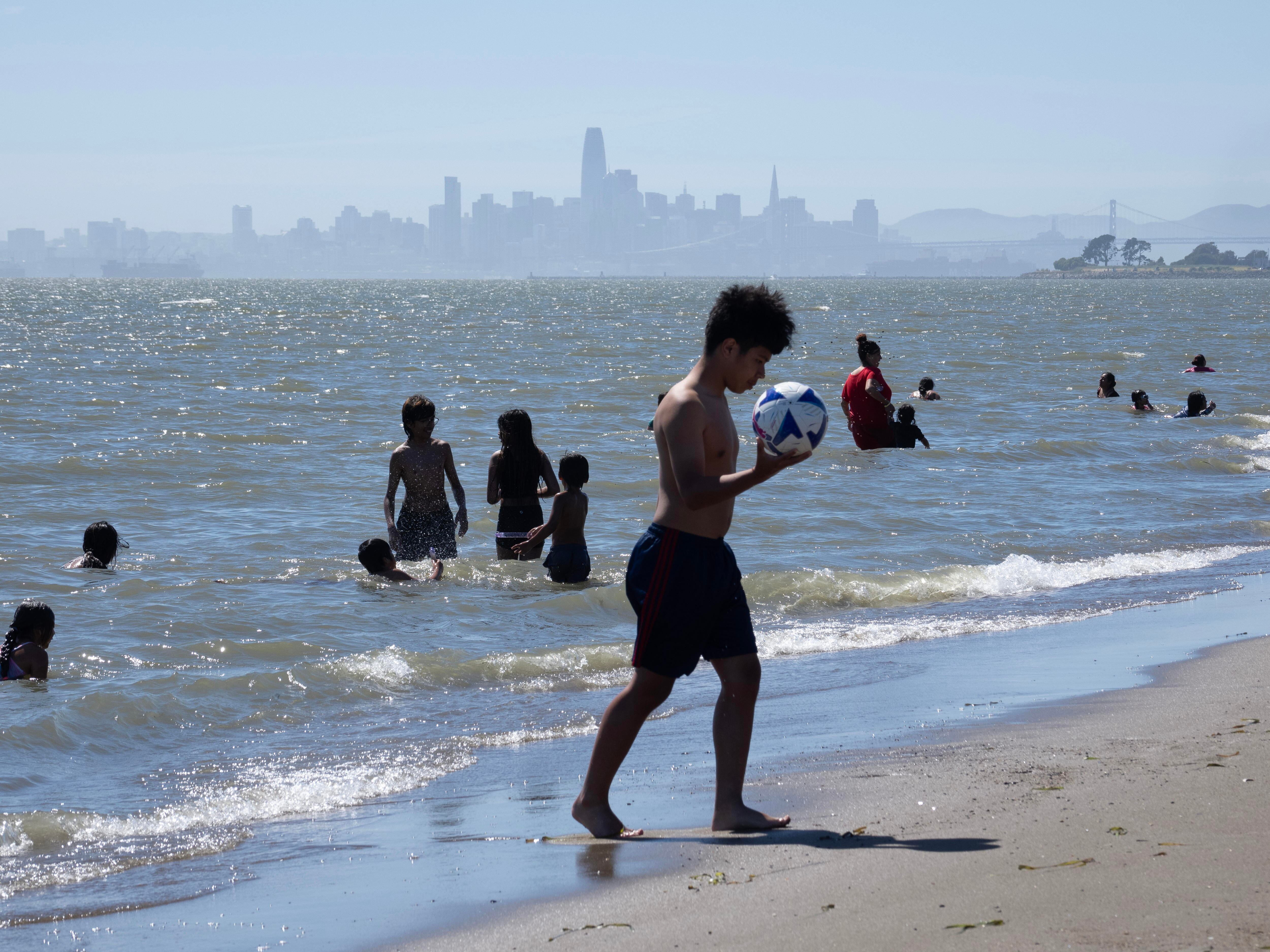 Un joven juega con un balón de fútbol mientras bañistas se refrescan frente a la playa de Alameda, California (EE UU), el 5 de junio 2024. 