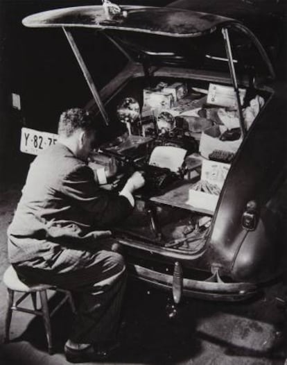 Weegee sentado en un taburete escribe a máquina en el maletero de su Chevrolet mientras fuma y se ilumina con una linterna, en 1942.