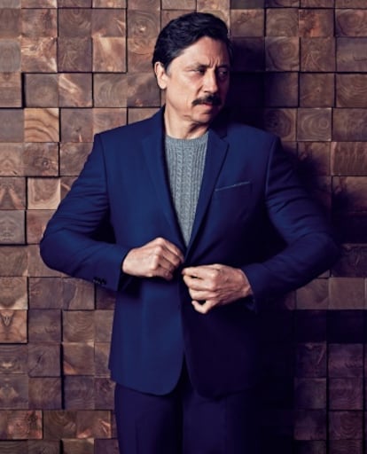 Carlos Bardem posa para ICON con traje azul y jersey, los dos, de Mango Man. Tras él, un panel de madera recuperada en desastres naturales de Wonderwall Studios para NorthView.