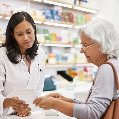 Senior customer showing prescription to female doctor. Cashier is assisting elderly woman at checkout counter. They are standing at pharmacy.