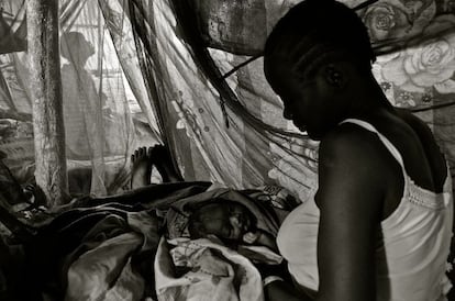 Una madre sostiene a su recién nacido en el campo de tránsito. El bebé nació a primera hora de la madrugada, sobre las 2, y por la tarde aún no había conseguido darle el pecho. Nuestro equipo la visitó en la tienda donde se encontraba y decidió trasladarla a Dzaipi para que fuera examinada por una matrona y un doctor de Médicos Sin Fronteras. En la imagen se puede distinguir la mosquitera, que muchas veces se utiliza mal porque no se cuelga correctamente o porque se usa como material de construcción de las casas. También se puede distinguir a una persona al fondo de la imagen, que demuestra la falta de privacidad inherente a este tipo de asentamientos. Teniendo en cuenta que son espacios cerrados, es fácil suponer que la higiene puede ser un problema, especialmente si se produce un parto. ¿Dónde se tira la placenta? ¿Quién limpia la sangre y los utensilios utilizados en el alumbramiento?