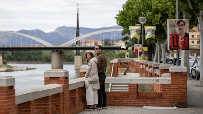 Una pareja observa el río Ebro en Tortosa, junto a carteles electorales con las imágenes de Salvador Illa o Pere Aragonès.