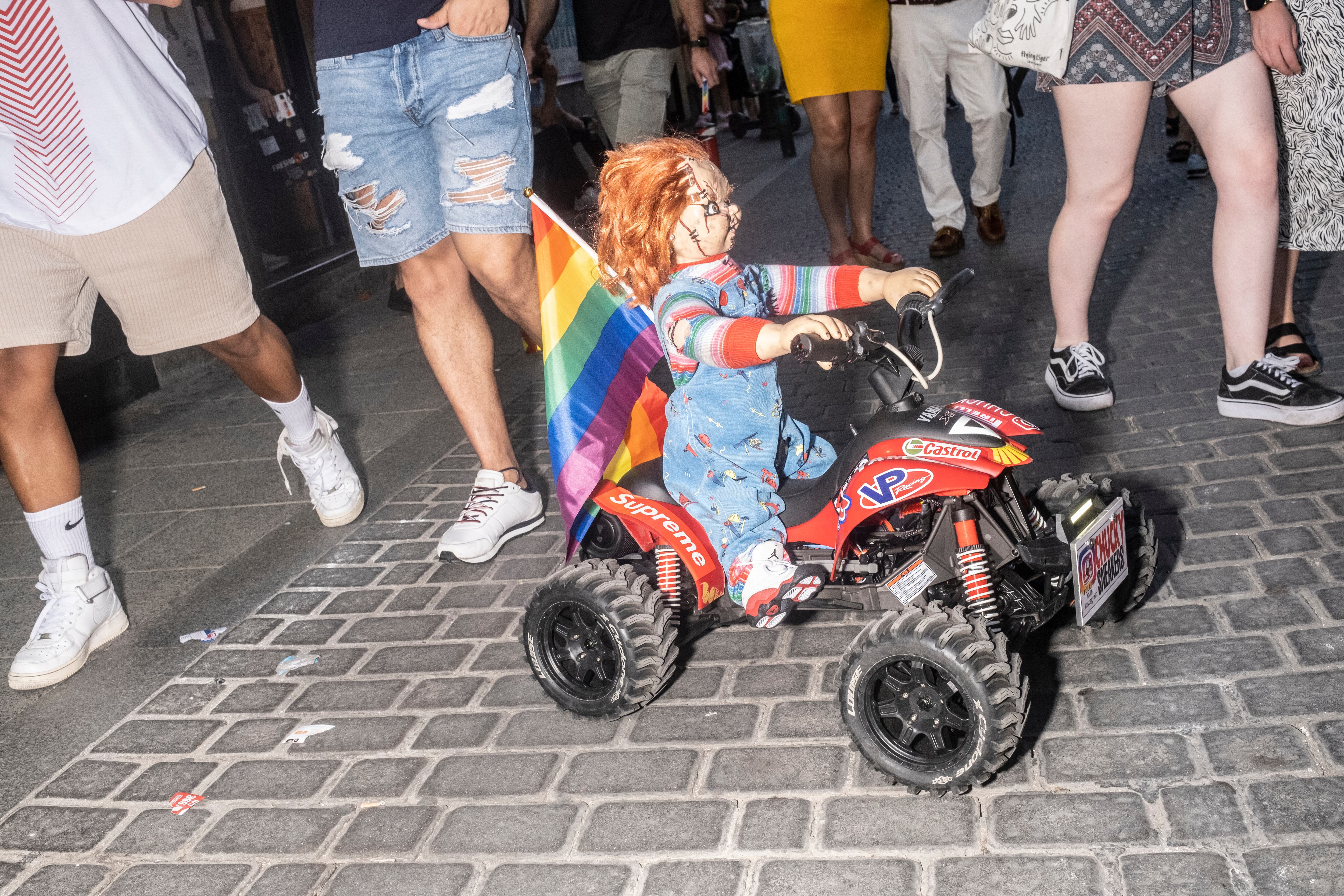 Un coche teledirigido con el muñeco Chucky y la bandera del orgullo, por una de las calles de Chueca.