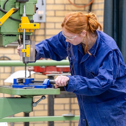 Two individuals in blue coveralls are working with a green drill press in an industrial setting, with one operating the machine and the other observing and taking notes