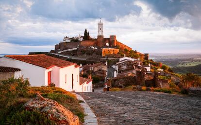 Para muchos, Monsaraz es uno de los pueblos más bellos de Portugal y <a href="https://elviajero.elpais.com/elviajero/2019/10/24/actualidad/1571905161_452130.html" target="_blank">uno de los más representativos del Alentejo</a>. Esta aldea diminuta encalada irrumpe en lo alto de un cerro coronado por un castillo, con vistas panorámicas de la presa de Alqueva y un paisaje salpicado de olivos. Está solo a 30 kilómetros de Villanueva del Fresno (Badajoz) y hoy vive del turismo, con un puñado de restaurantes, pensiones y tiendas artesanales. Merece la pena visitarla para saborear el Portugal más tradicional, recorrer sus calles soñolientas y probar la cocina alentejana. El mejor momento es cuando se despierta por la mañana, o en la tranquilidad de un anochecer de invierno. El paisaje que rodea Monsaraz está repleto de megalitos neolíticos que pueden descubrirse mientras paseamos entre olivares y campos en flor (están señalizados, pero encontrarlos es emocionante). El más espectacular es el Cromeleque do Xerez, un conjunto presidido por un triunfal menhir de siete toneladas. Las rocas se hallaban cinco kilómetros al sur de Monsaraz, pero fueron trasladadas para salvarlas de la inundación de la enorme presa de Barragem do Alqueva.