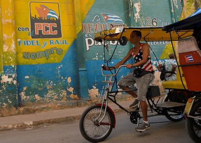 Un hombre vestido con una camiseta con la bandera de Estados Unidos pasa en su bicitaxi frente a un mural del Partido Comunista Cubano.