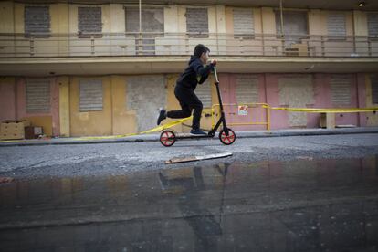 Un niño pasa con su patinete delante de las vallas de la calle de San Fulgencio, mientras se refleja en un charco. Al fondo, aparecen las viviendas con ventanas y puertas tapiadas.