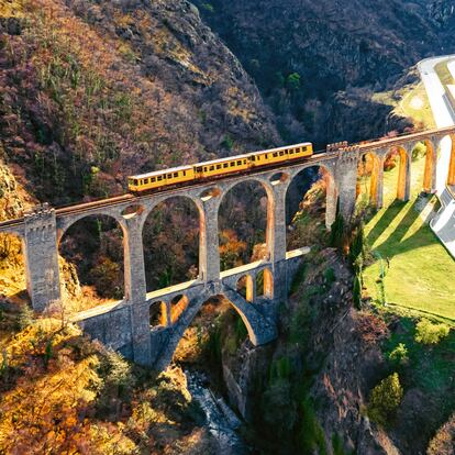 The Train Jaune in the Catalan region of the Pyrenees Orientales in France, crossing old arch bridge made with cool arches, with stunning valley, dramatic landscape with rain and sun. Fontpédrouse. France.