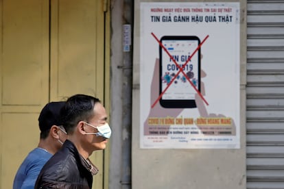 Dos hombres protegidos con mascarillas pasan frente a un cartel en Hanoi (Vietnam) que advierte contra la difusión de bulos.