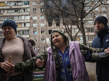 People help an elderly woman to walk in a street with an apartment building hit by shelling in the background in Mariupol, Ukraine, Monday, March 7, 2022. (AP Photo/Evgeniy Maloletka)