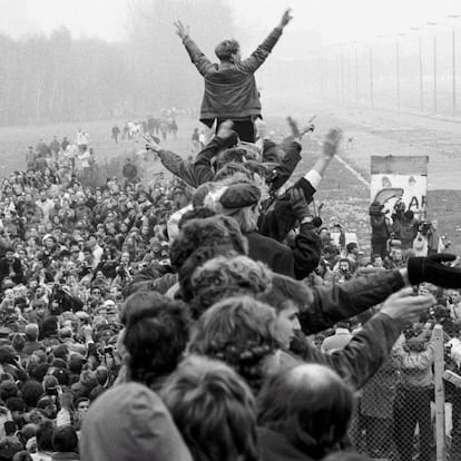 Entre una multitud encaramada al muro de Berlín, un joven en pie levanta los brazos y hace el signo de la victoria con ambas manos. A la izquierda se observa la plaza de Postdamer repleta de gente.