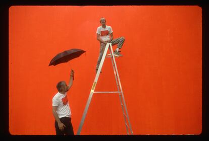 Keith Haring, en 1987, sobre una escalera frente al muro sobre el que va a pintar su famoso mural 'Crack is Wack'. A sus pies, sostiene un paraguas el entonces responsable de parques y jardines de Nueva York, Stern, que lleva una camiseta con el dibujo de Haring. | 