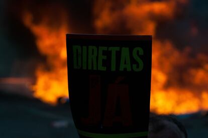 Manifestante pede eleições antecipadas diretas, em Brasília. Ao fundo, barricada com fogo na Esplanada dos Ministérios. 